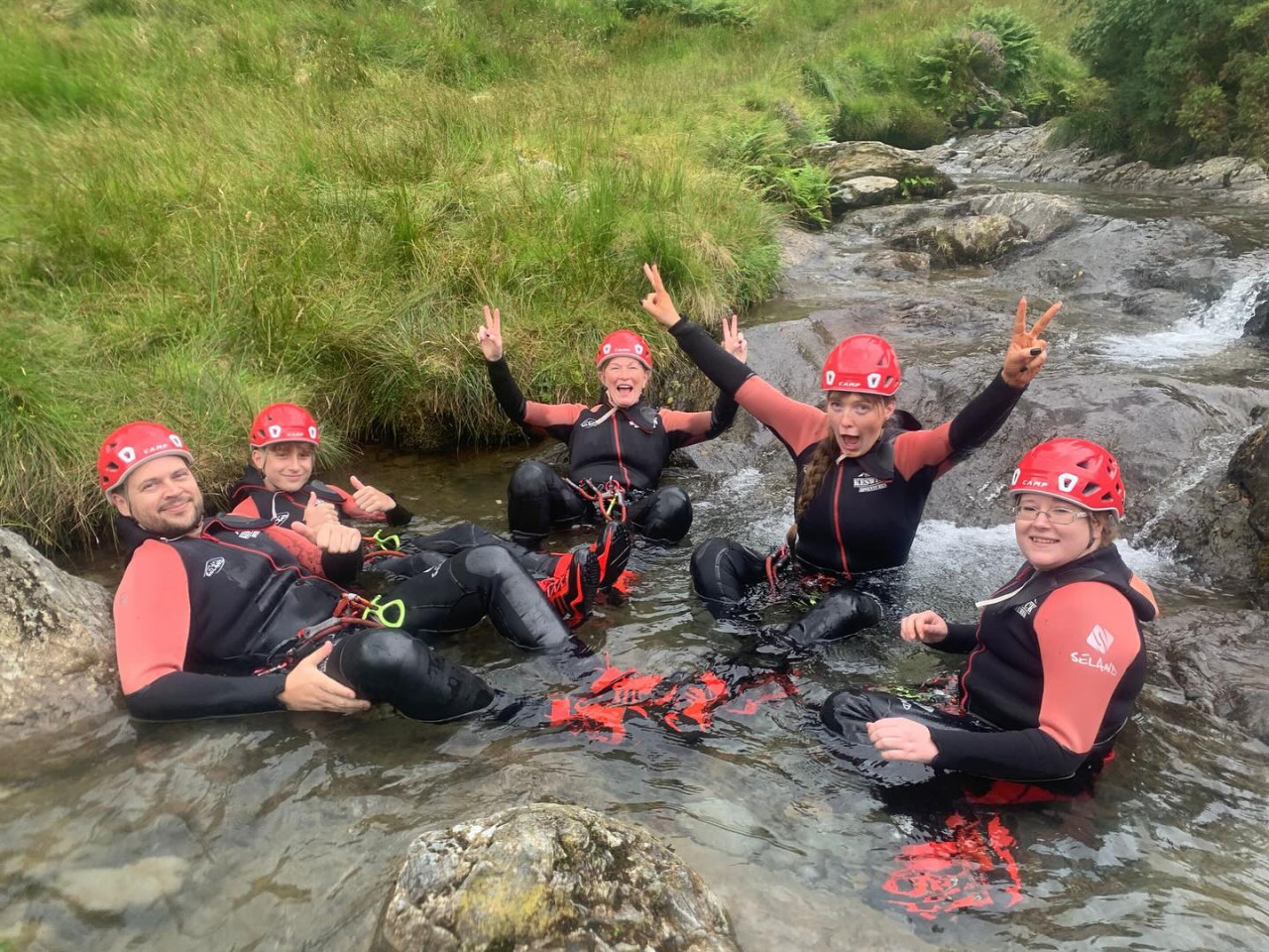 Family of 5 people in a shallow river, showing thumbs up and peace signs